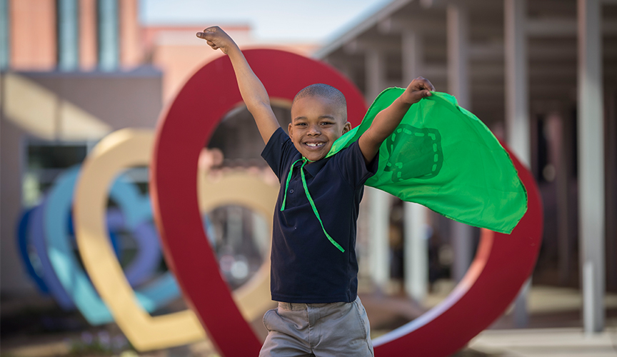 Child with superhero cape stands in front of a Le Bonheur heart.